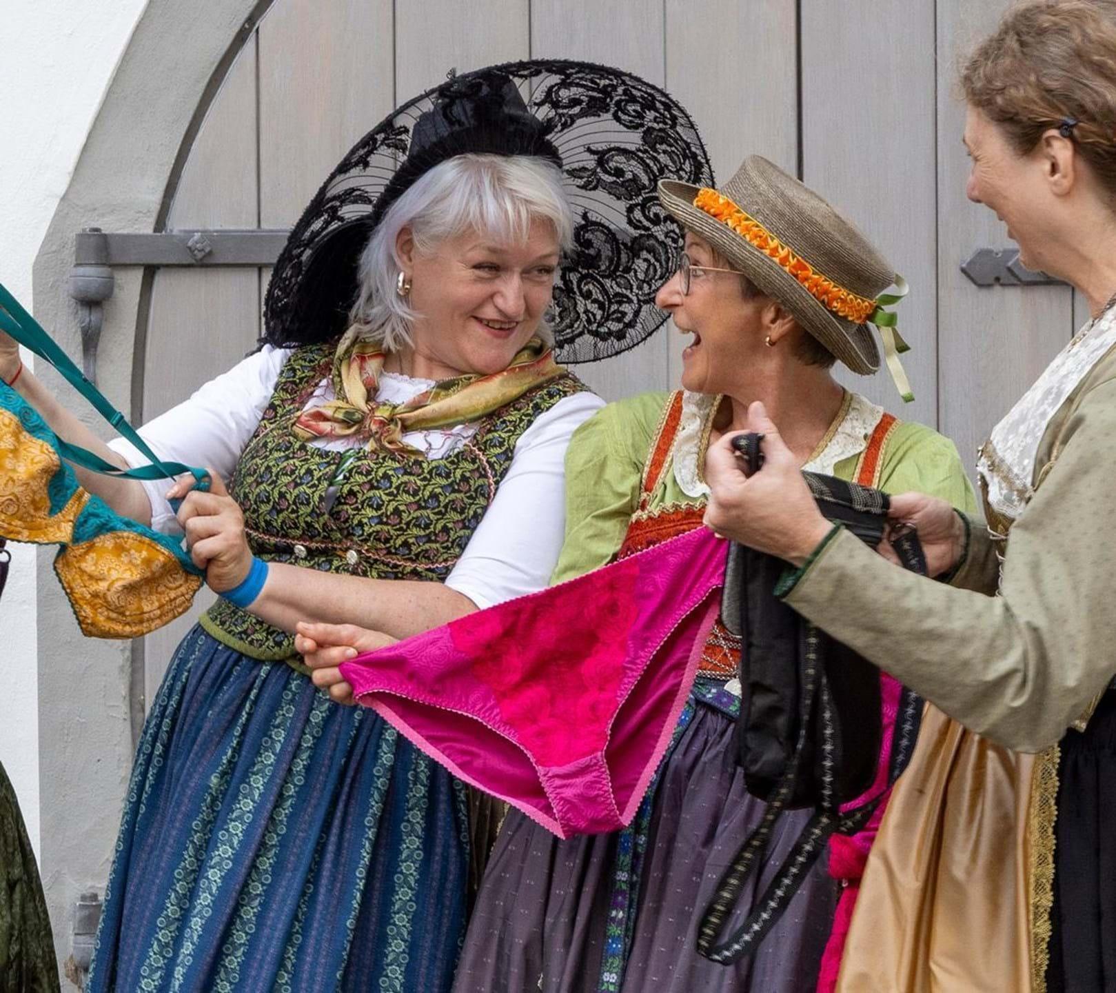 Drei Frauen in Tracht betrachten Unterwäsche und lachen dabei